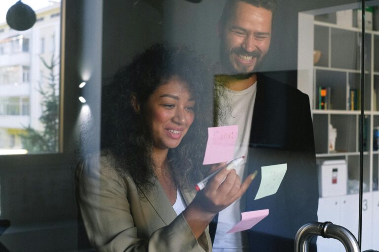 A woman and a man smiling and brainstorming content strategy ideas with colorful post-it notes in front of a whiteboard.